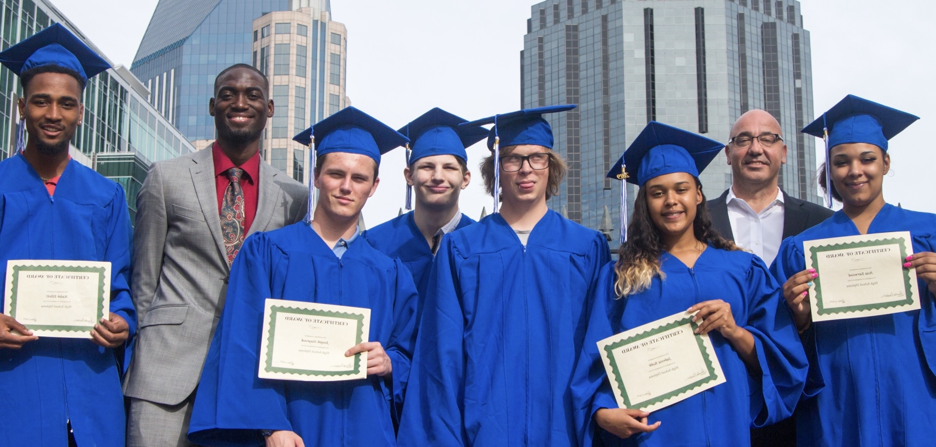 Six diverse graduates in blue caps and gowns holding certificates, standing with two adults in business attire in an urban setting.