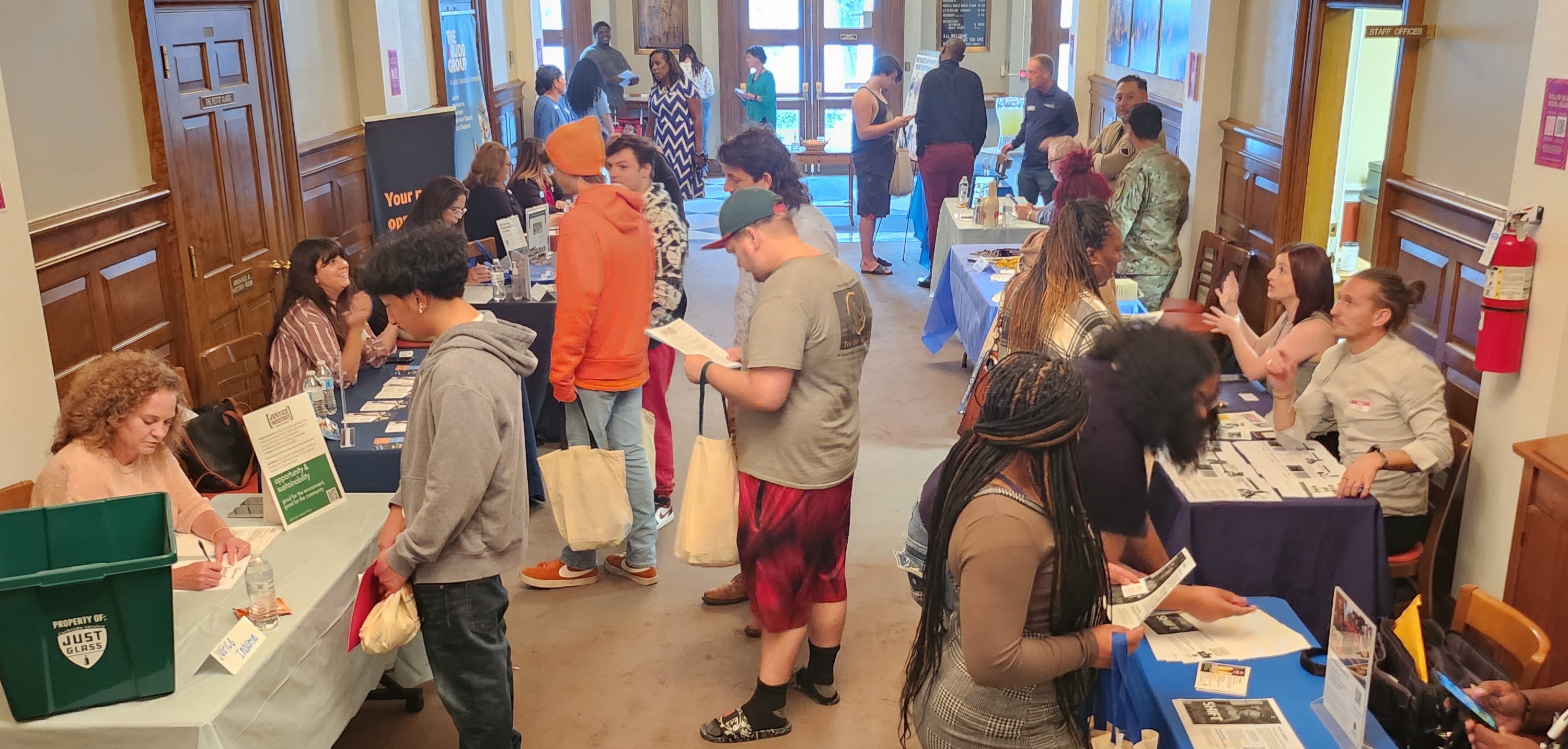 People interacting at various booths in a hallway during an indoor community event or job fair.
