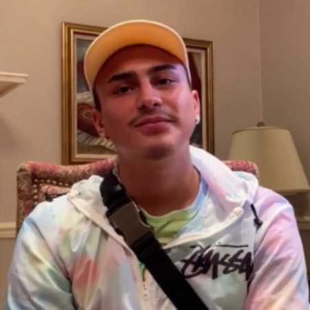Young man wearing a light-colored hoodie with a black strap across his chest and a yellow baseball cap, sitting in a room with a framed painting and a table lamp.