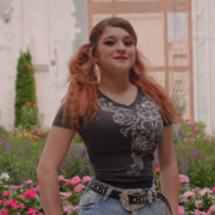 Young woman with long reddish hair in pigtails, wearing a black patterned t-shirt and light blue jeans, standing in a garden with colorful flowers.