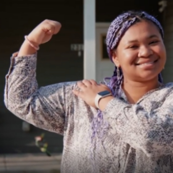 Woman with purple braided hair smiling and flexing her arm muscle outdoors.
