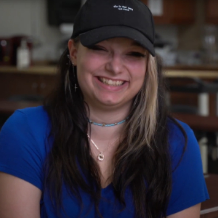 Young woman with long dark hair and a blonde streak, wearing a black cap and blue shirt, smiling warmly.