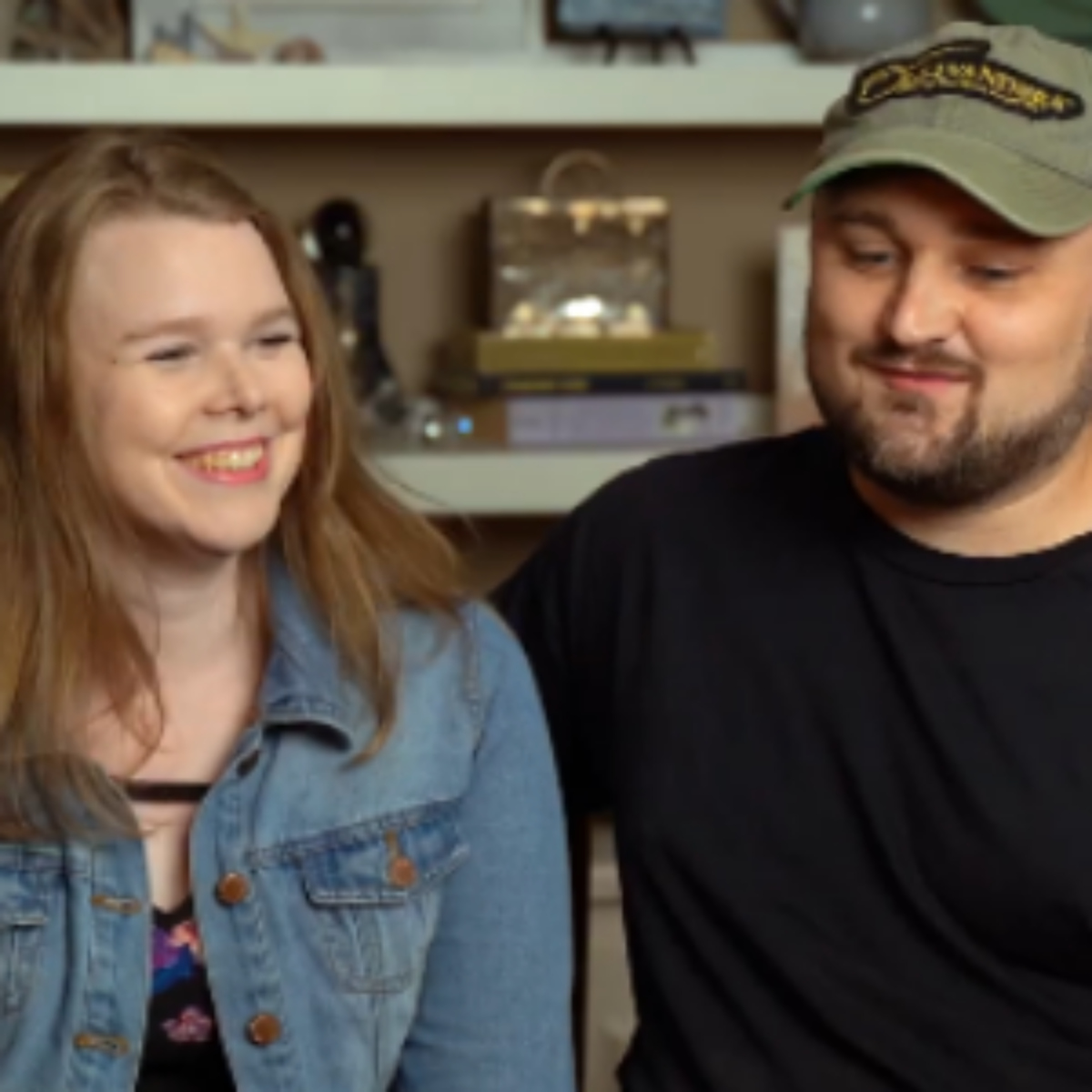 Smiling young woman in a denim jacket and bearded man in a baseball cap sitting together indoors.