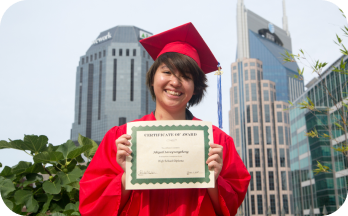Smiling graduate in red cap and gown holding a certificate outdoors with tall buildings in the background.