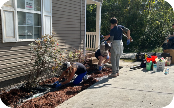 Three people working together to plant or garden in a narrow bed beside a house covered with brown mulch.