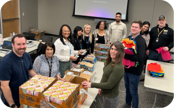 Group of people smiling and packing boxed food items on tables in a room.