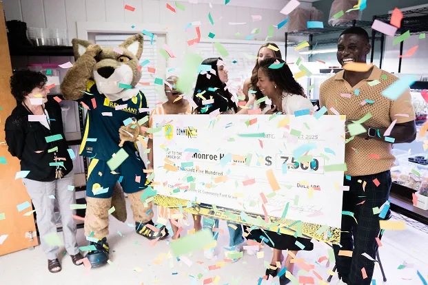 Group of people celebrating with a large check for $7,500 while colorful confetti falls around them in a bright room.