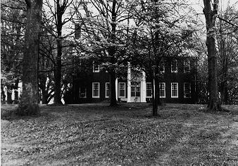 Black and white photo of a two-story brick house with columns in front, partially obscured by leafless trees on a grassy lawn.