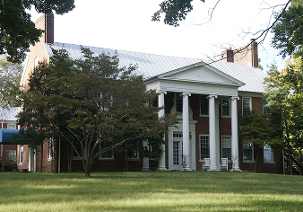 Historic brick building with white columns and a silver roof surrounded by trees and lawn.