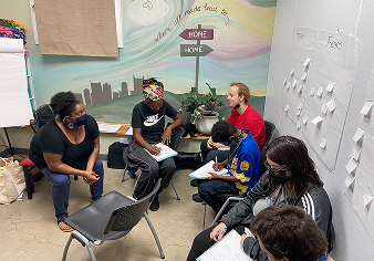 Six people sitting in a circle in a room, engaged in discussion and note-taking, with a colorful mural and sticky notes on the walls.