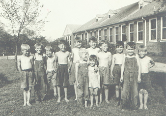 Black and white photo of twelve boys standing in a row outside a large building, most wearing overalls or shorts, some shirtless.