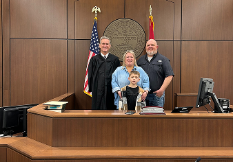 Judge standing behind a young boy at a courtroom bench with the boy's smiling parents and U.S. and state flags in the background.