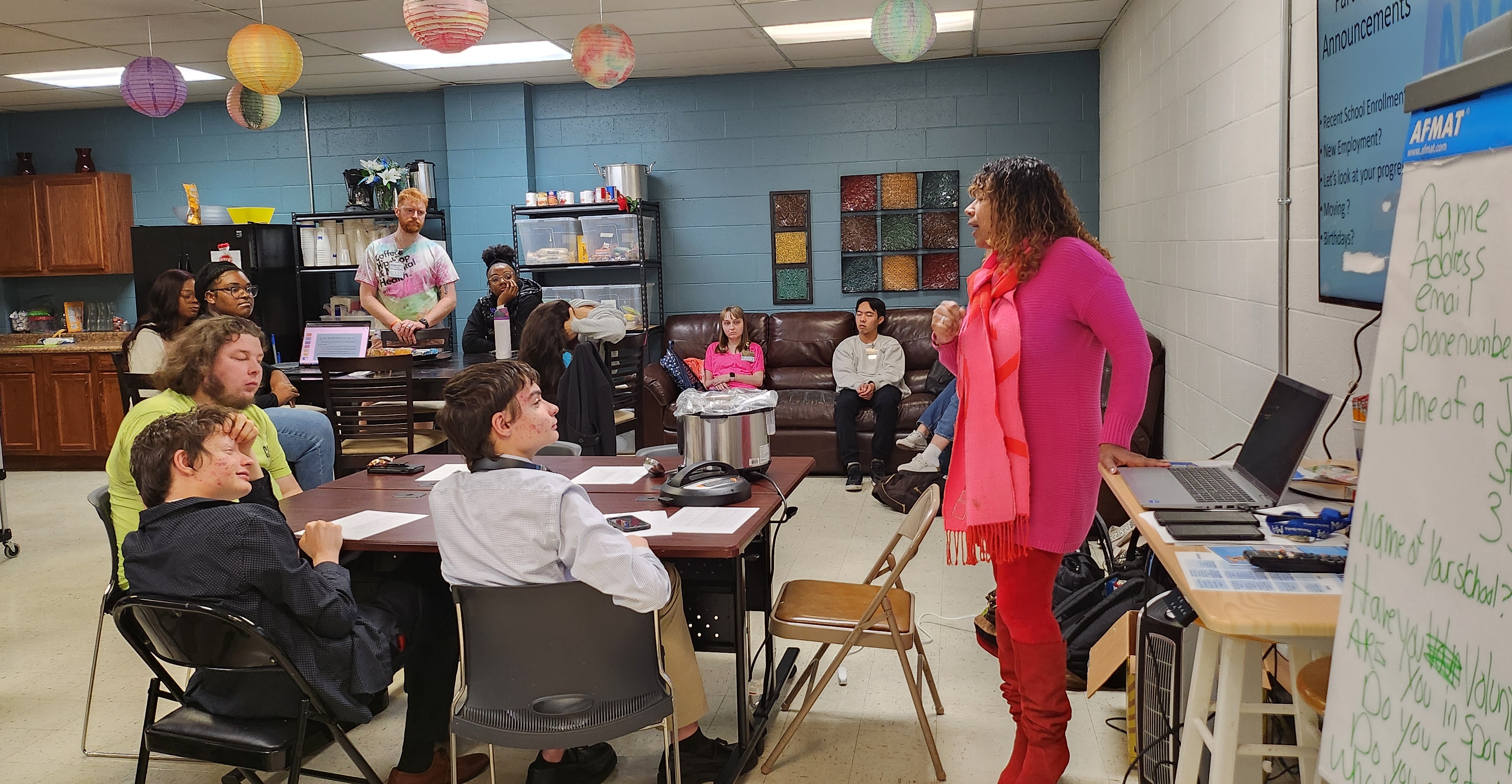 A woman in a pink sweater and red boots stands and speaks to a group of diverse people seated around tables and on a couch in a classroom or community room decorated with hanging paper lanterns.