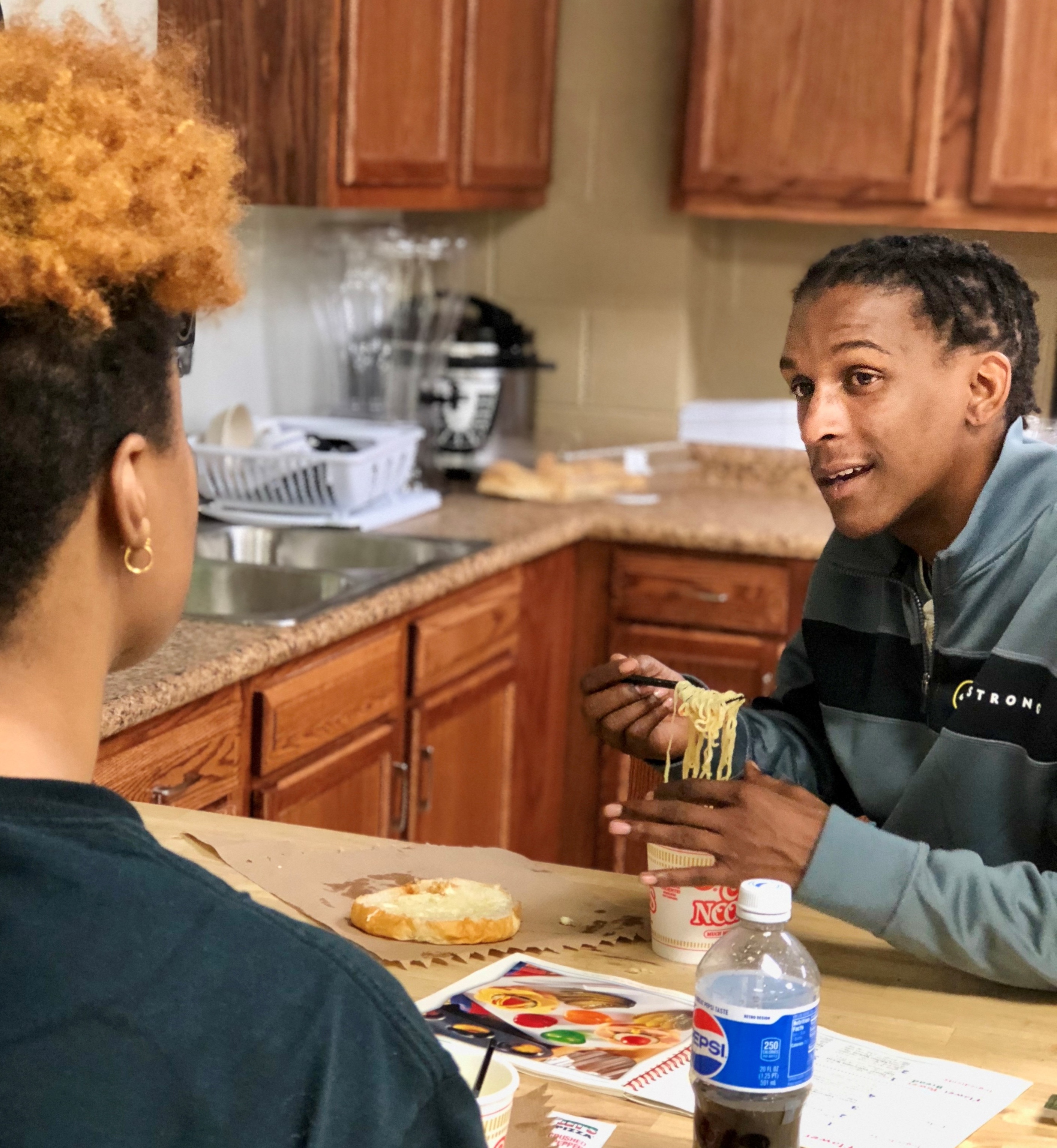 Two people sit at a kitchen table with wooden cabinets behind them; one eats noodles from a cup, the other faces away with a small round sandwich on the table, along with a bottle of Pepsi and a color palette book.