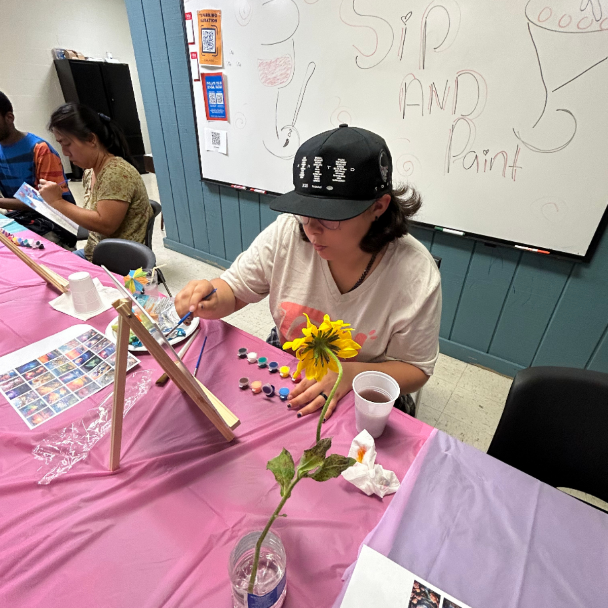 Person wearing glasses and a black cap painting on a canvas at a table with paint pots, a cup, and a yellow flower in a bottle; a sign behind reads Sip and Paint.