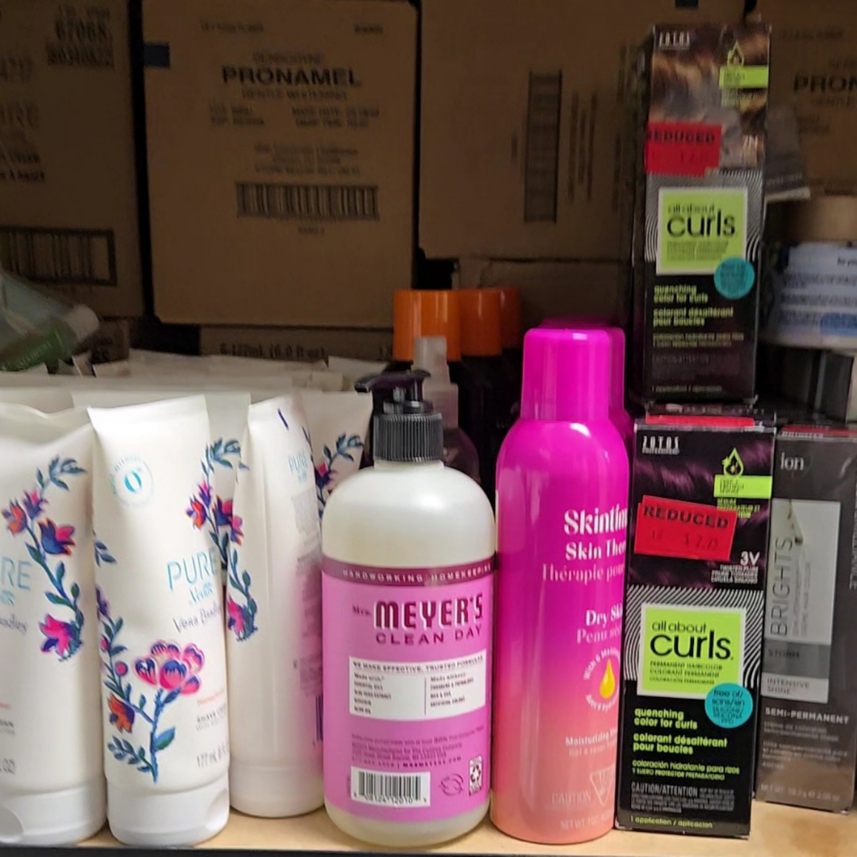 Shelf with personal care products including Mrs. Meyer's Clean Day lotion, Skintifique dry skin therapy, Pure products in white tubes with floral design, and All About Curls hair color boxes with reduced price stickers.