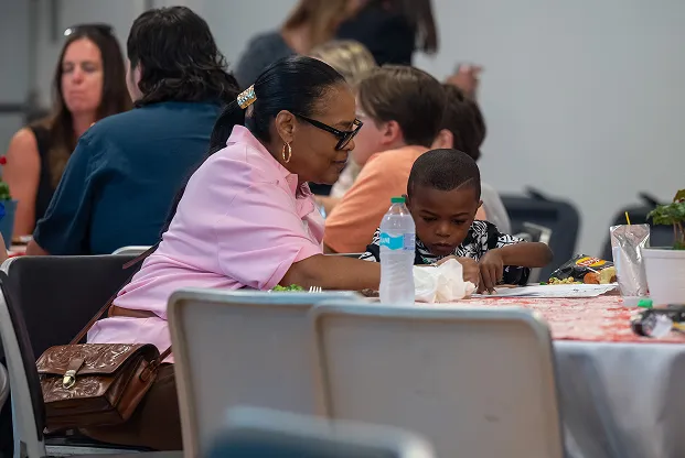 Woman in a pink shirt sitting at a table with a young boy, both focused on an activity on the table, with other people seated and talking in the background.
