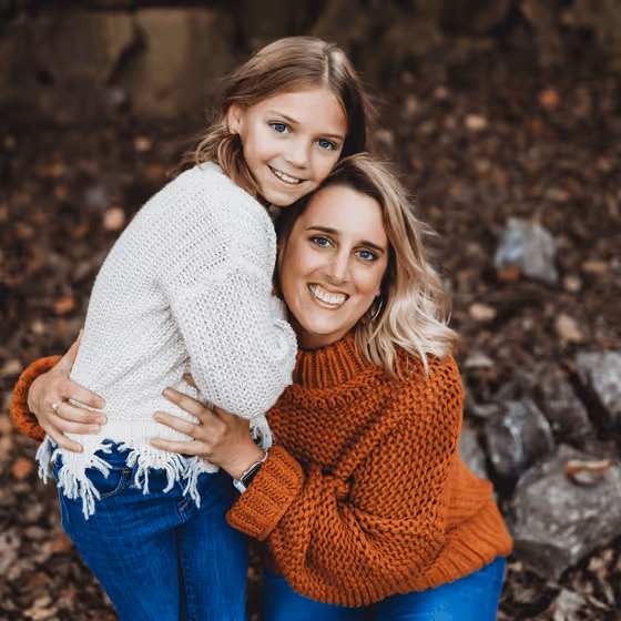 Smiling woman in orange sweater hugging a girl in white sweater outdoors with autumn leaves.