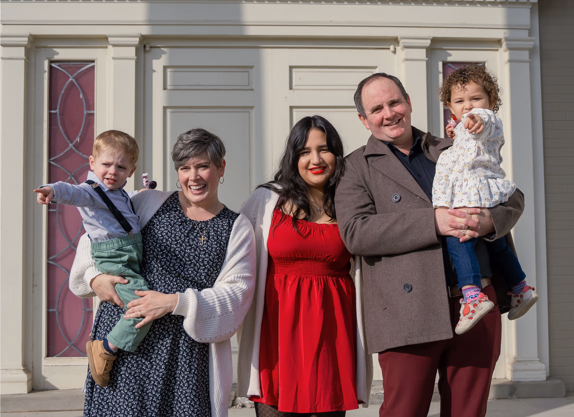 Two adults each holding a child, all smiling in front of a building with white and red doors.