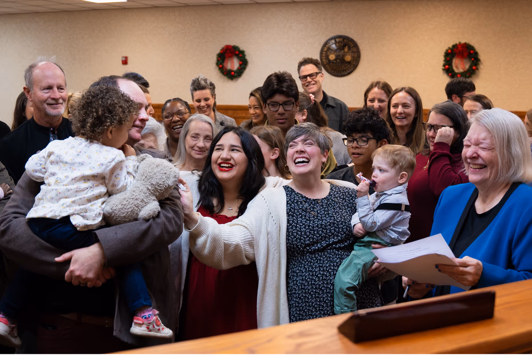 A foster family smiling and celebrating together indoors, with two children held by adults and holiday wreaths on the beige walls behind them.