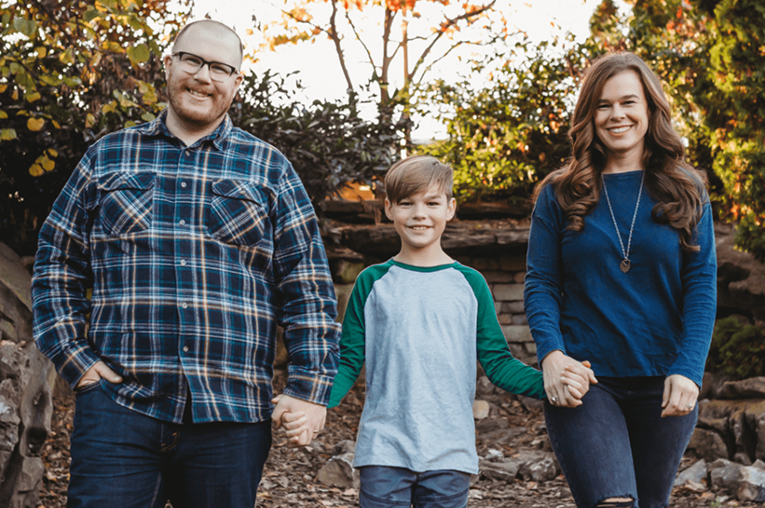 Smiling foster family showing a man, woman, and boy holding hands outdoors with trees and rocks in the background.