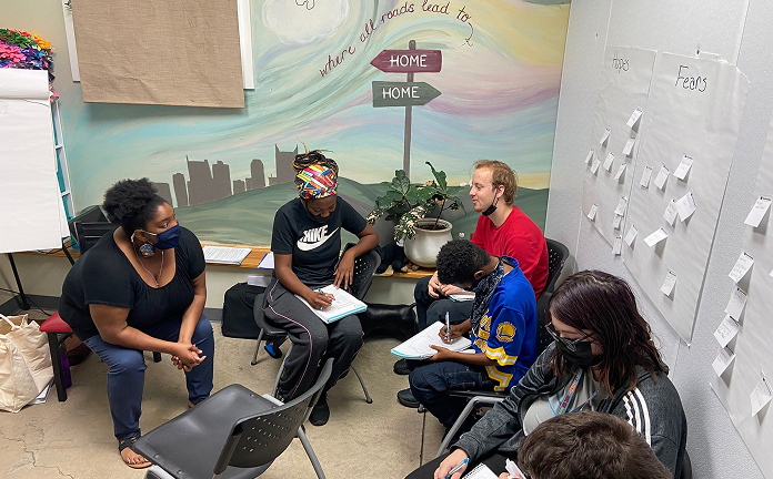 A group of six people sitting in a small room in a discussion; some are writing on notepads. The room has a mural with signs reading 'HOME' and posters labeled 'Hopes' and 'Fears' with sticky notes.