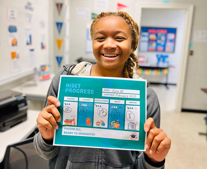 Smiling young woman holding a HISAT progress report showing passing scores in reading, science, social studies, math, and writing, with a note that says passed all and ready to graduate.