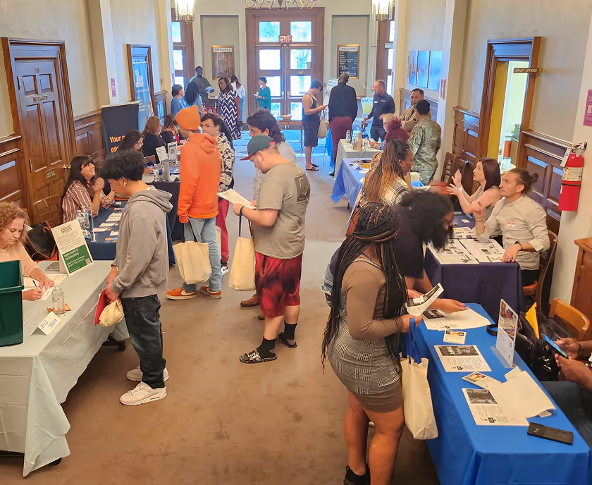 Indoor job fair with multiple booths on both sides of a hallway where diverse attendees engage with recruiters and review materials.