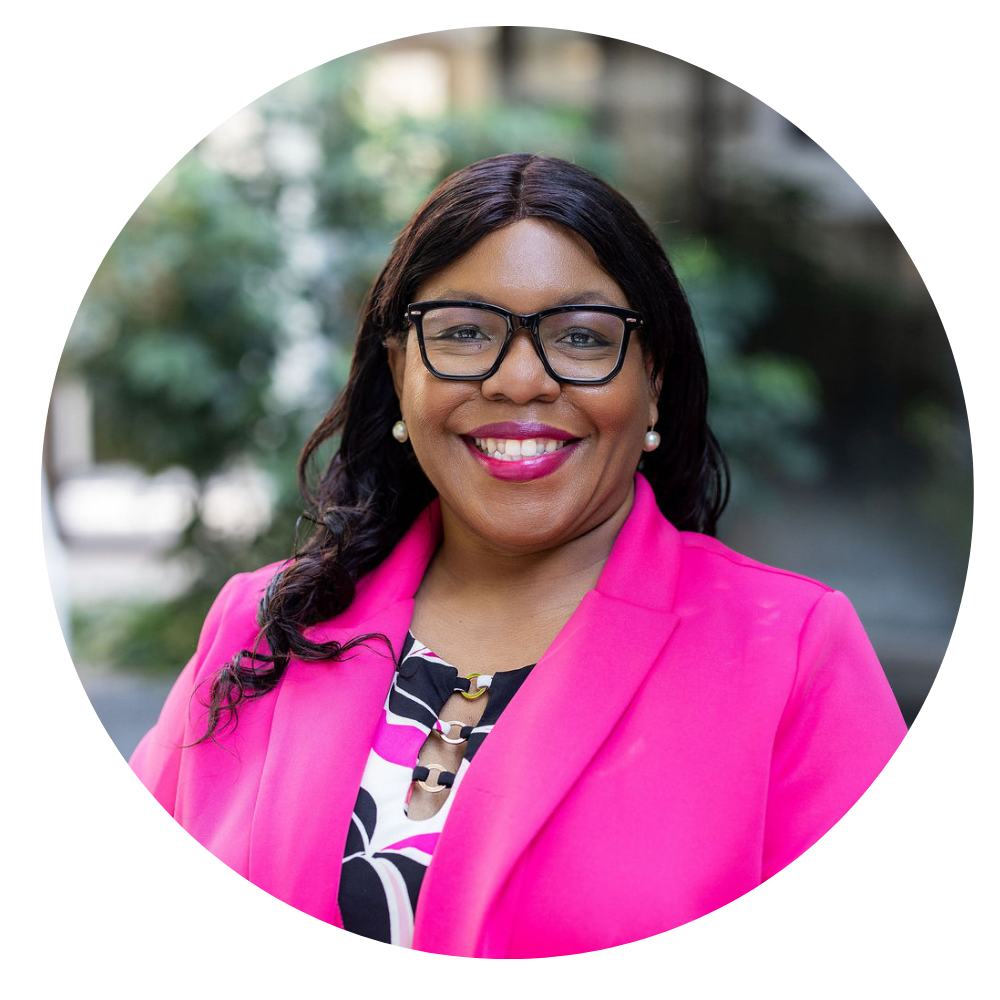 Shan Edmondson, Sr. Director of Foster Care. Photo shows a Smiling woman with dark hair wearing black framed glasses, pearl earrings, a bright pink blazer, and a patterned top, posed outdoors with blurred greenery in the background.