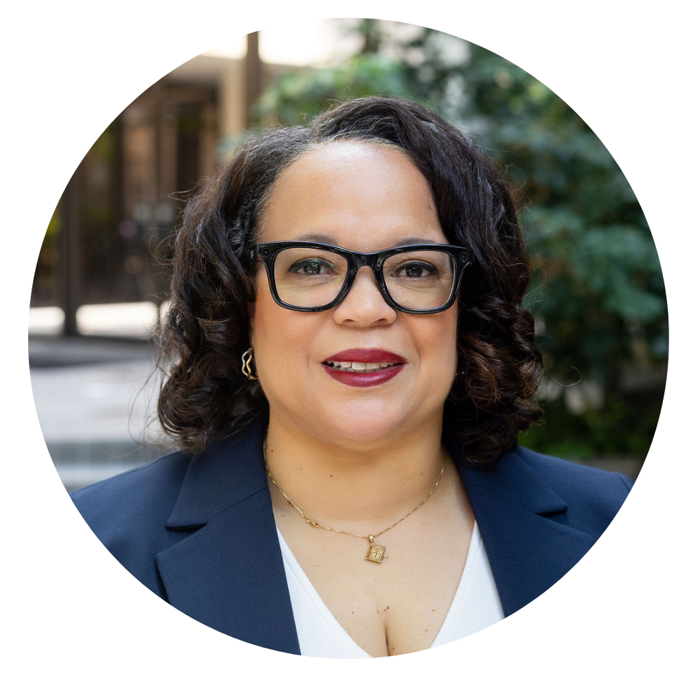 Quatrece McKinney, Vice President of Programs. Photo shows a Smiling woman with curly dark hair wearing black rectangular glasses, a navy blazer, white top, gold earrings, and a gold necklace with a square pendant, against a blurred outdoor background with greenery.