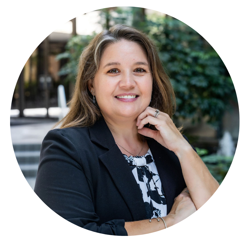 Deidre Matthews, Vice President of Finance & Administration. Photo shows a Smiling woman with long light brown hair wearing a black blazer over a patterned black and white blouse, posing outdoors with greenery in the background.