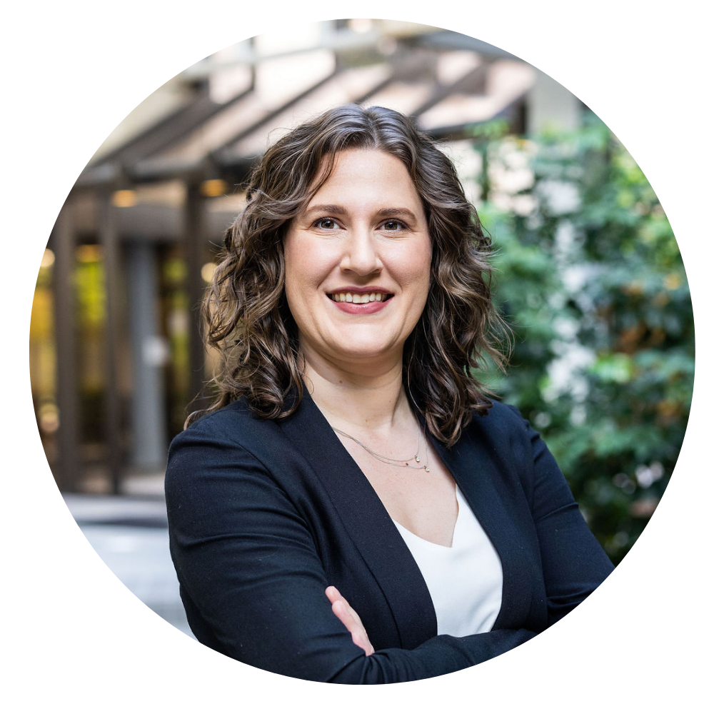 Kristen Heritage, Director of Data & Quality Assurance. Photo shows a Smiling woman with wavy brown hair wearing a black blazer and white top standing with crossed arms against a blurred outdoor background with green foliage and building elements.
