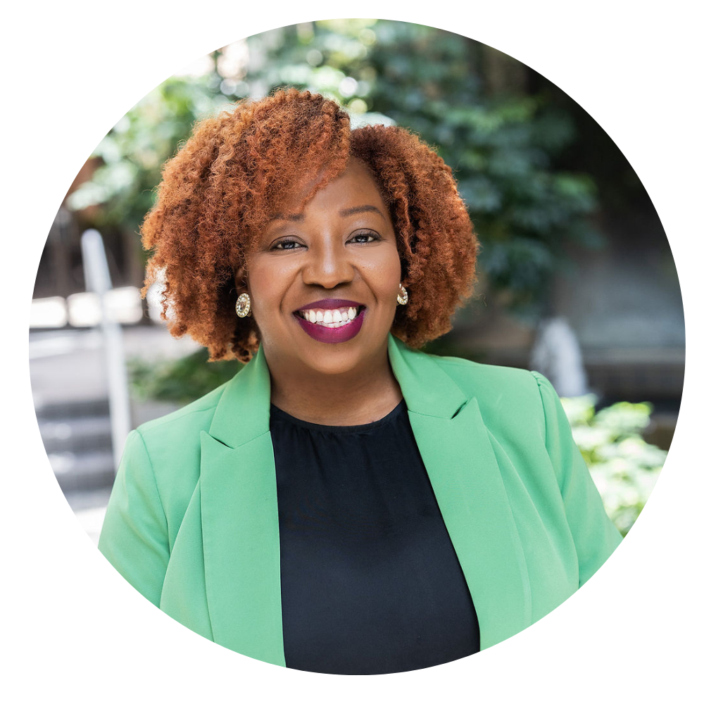 Sharica S. Ware, CAE, Senior Director of Development & Communications. Photo shows a smiling woman with curly reddish hair wearing a green blazer and black top, standing outdoors with blurred greenery in the background.