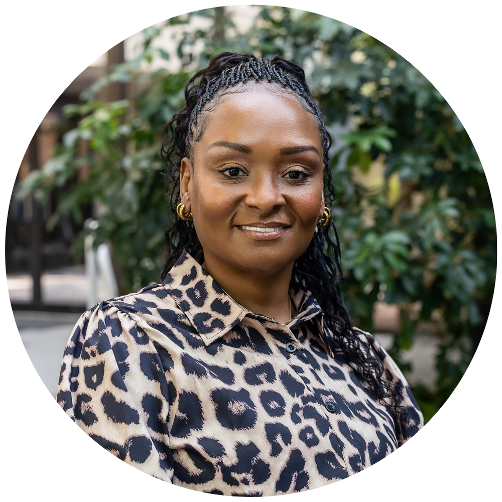 Headshot of LaKesha Malone. Smiling woman with braided hair pulled back, wearing gold hoop earrings and a leopard print blouse, standing outdoors with green foliage in the background.