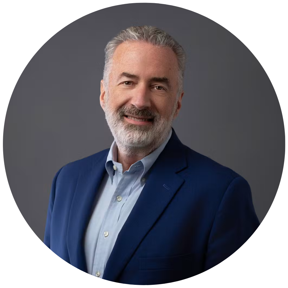 Headshot for Mark Patterson, a Smiling middle-aged man with gray hair and beard wearing a blue blazer and light blue shirt against a gray background.