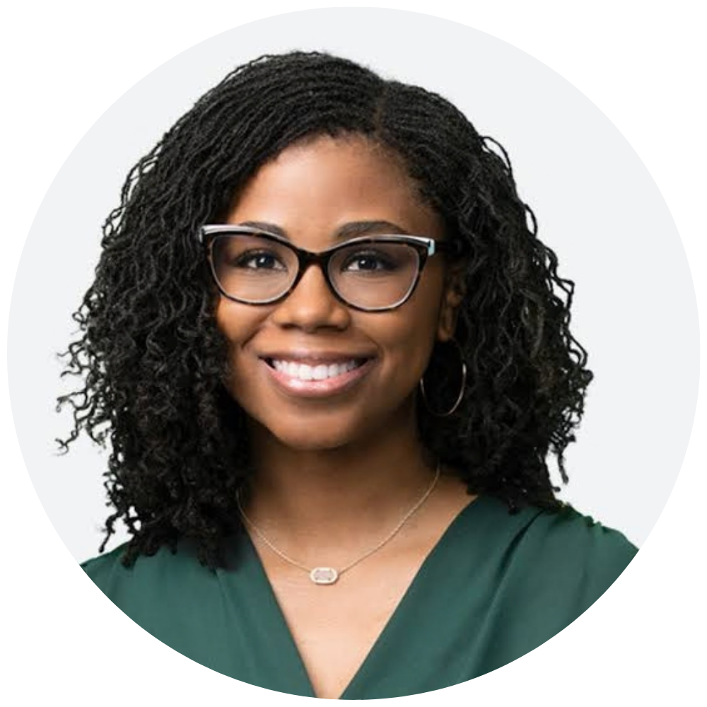 Smiling woman with curly hair wearing black glasses, hoop earrings, a green top, and a silver necklace against a plain light background.
