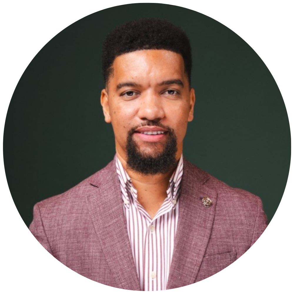 Headshot for Krishna Campbell. A man with short curly black hair and a goatee wearing a pink textured blazer over a white and pink striped shirt, posing in front of a dark green background.