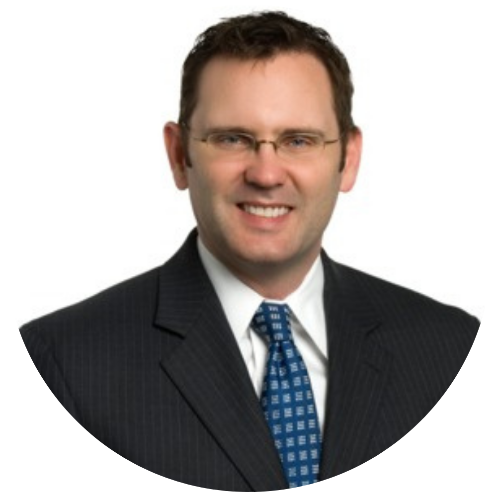 Portrait of a man with short brown hair and glasses wearing a dark pinstripe suit, white shirt, and blue patterned tie, smiling against a white background.