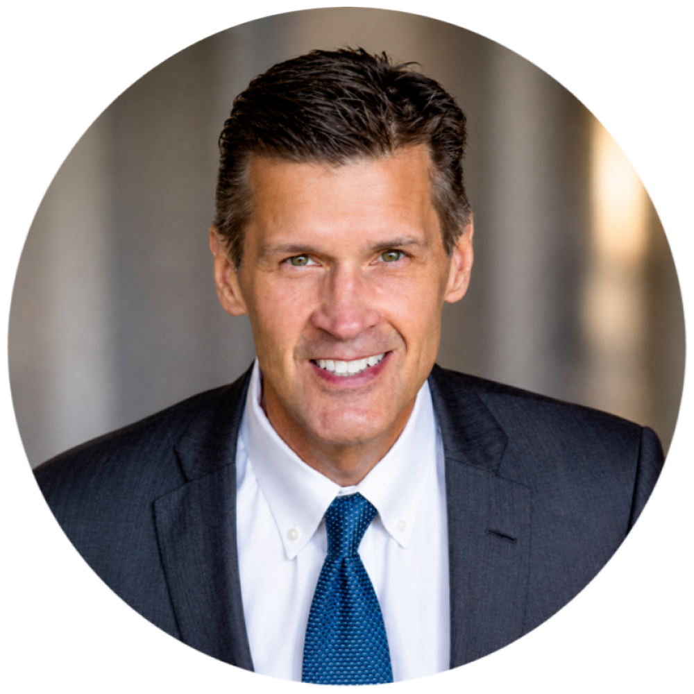 Portrait of a smiling man with short dark hair wearing a dark suit jacket, white dress shirt, and blue tie, set against a softly blurred background.