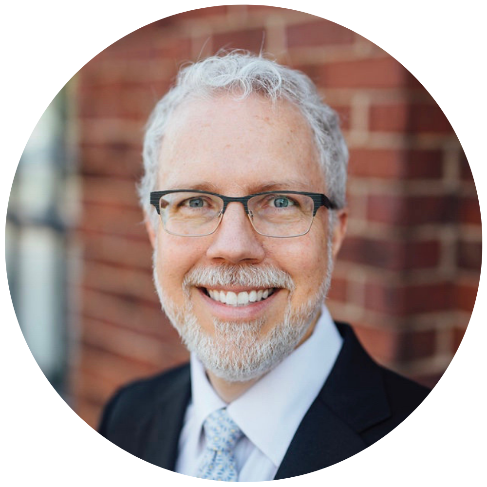 Smiling middle-aged man with gray hair, beard, and glasses wearing a black suit jacket, white shirt, and patterned tie, standing in front of a brick wall.