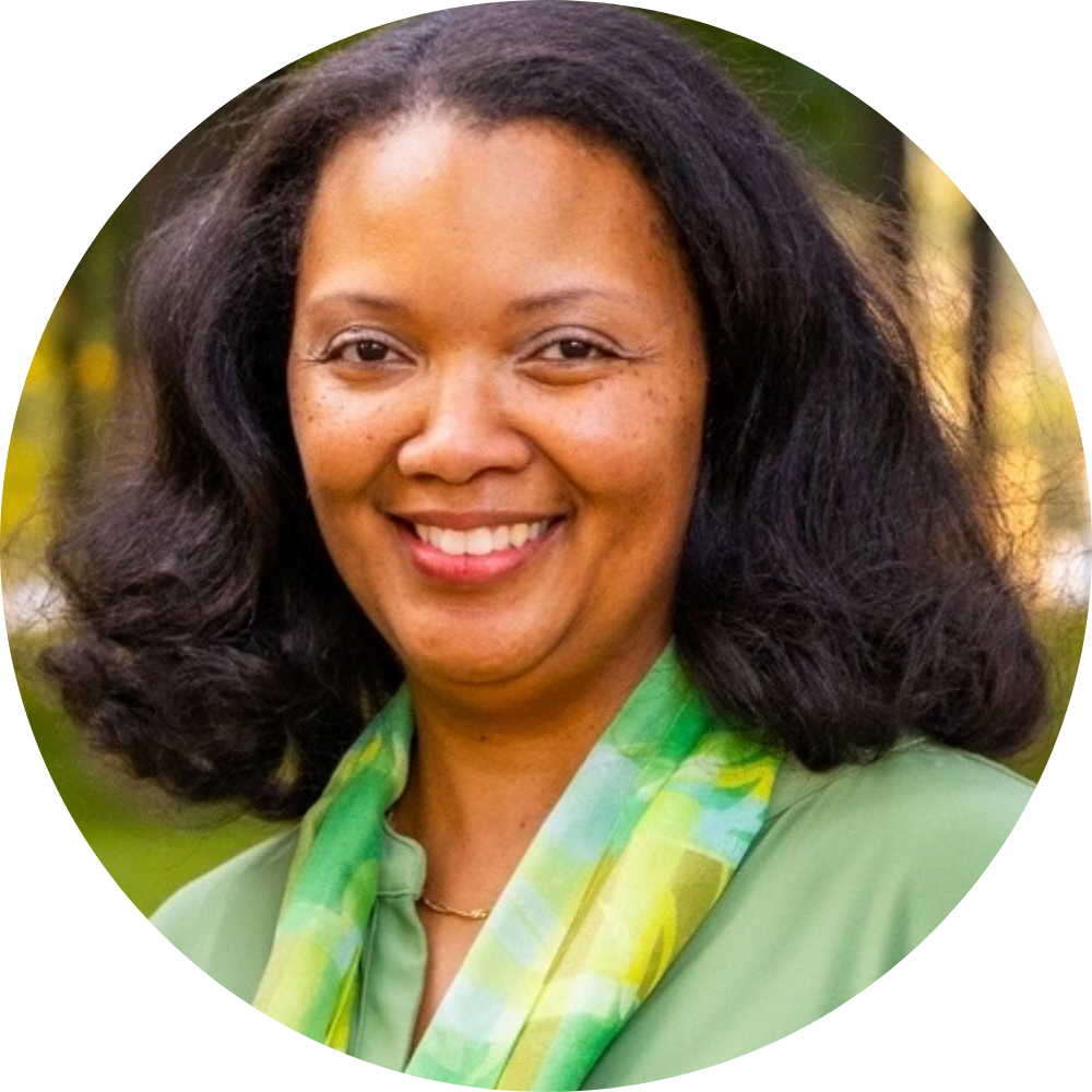 Headshot for Dena Cheatham. Shows a smiling woman with medium-dark skin, shoulder-length curly black hair, wearing a light green blouse and a green-yellow patterned scarf, outdoors with blurred trees in the background.