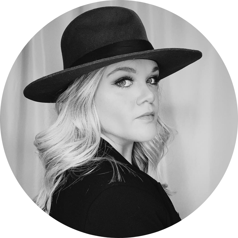 Black and white portrait of a woman with long wavy blonde hair wearing a wide-brimmed hat and a dark jacket, looking sideways toward the camera against a plain background.