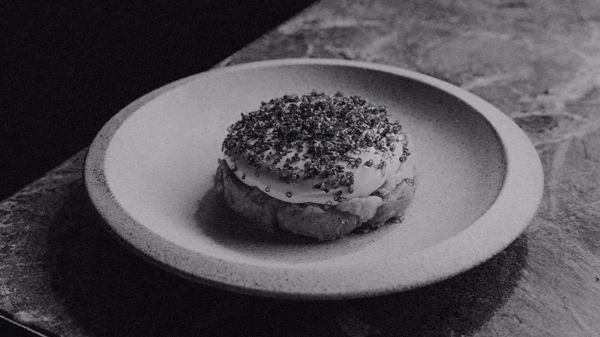 A round biscuit topped with creamy icing and sprinkled with small seeds, served on a plain ceramic plate on a wooden surface.