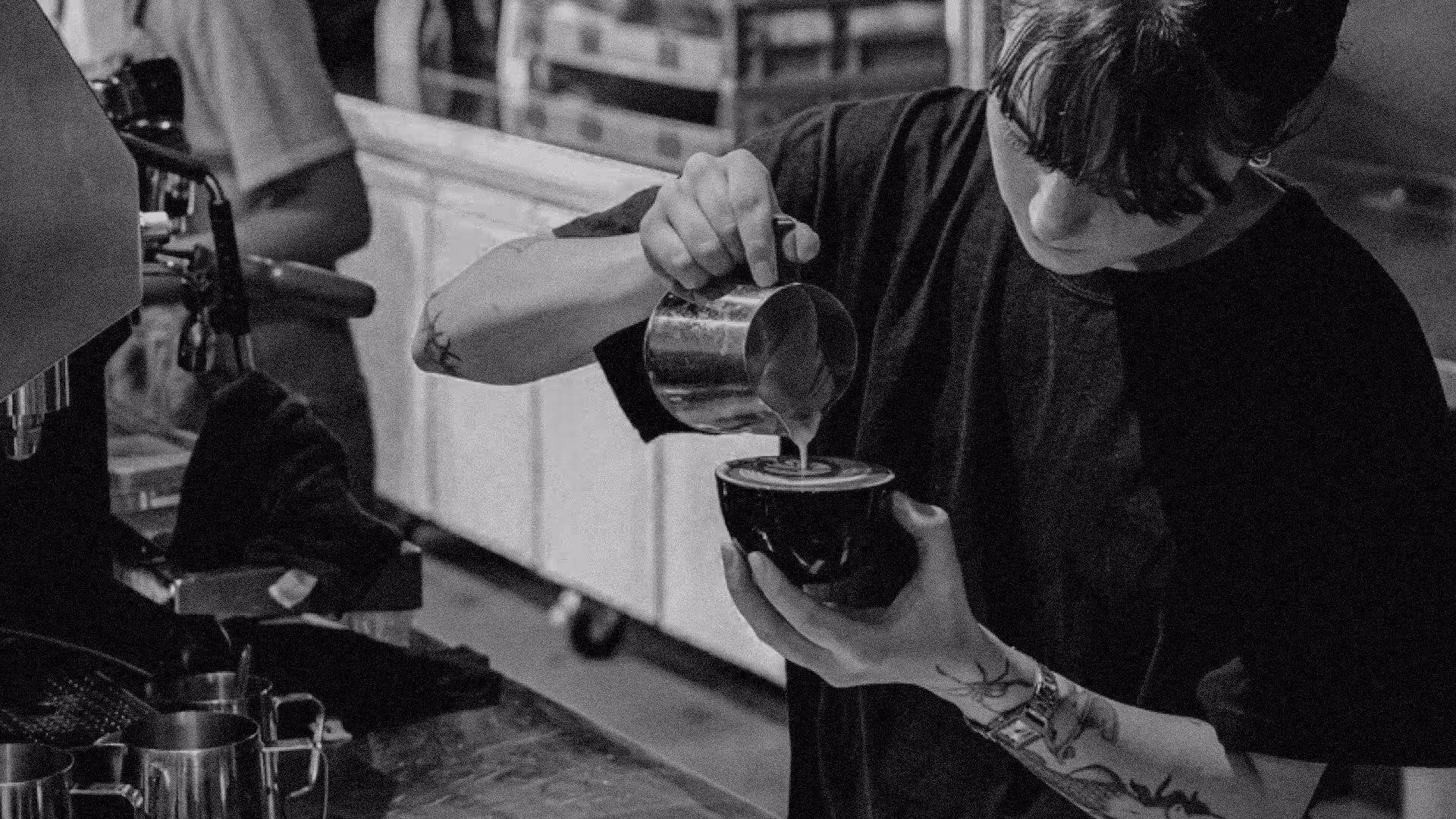 Barista with tattoos pouring steamed milk into a black cup to make latte art in a coffee shop.