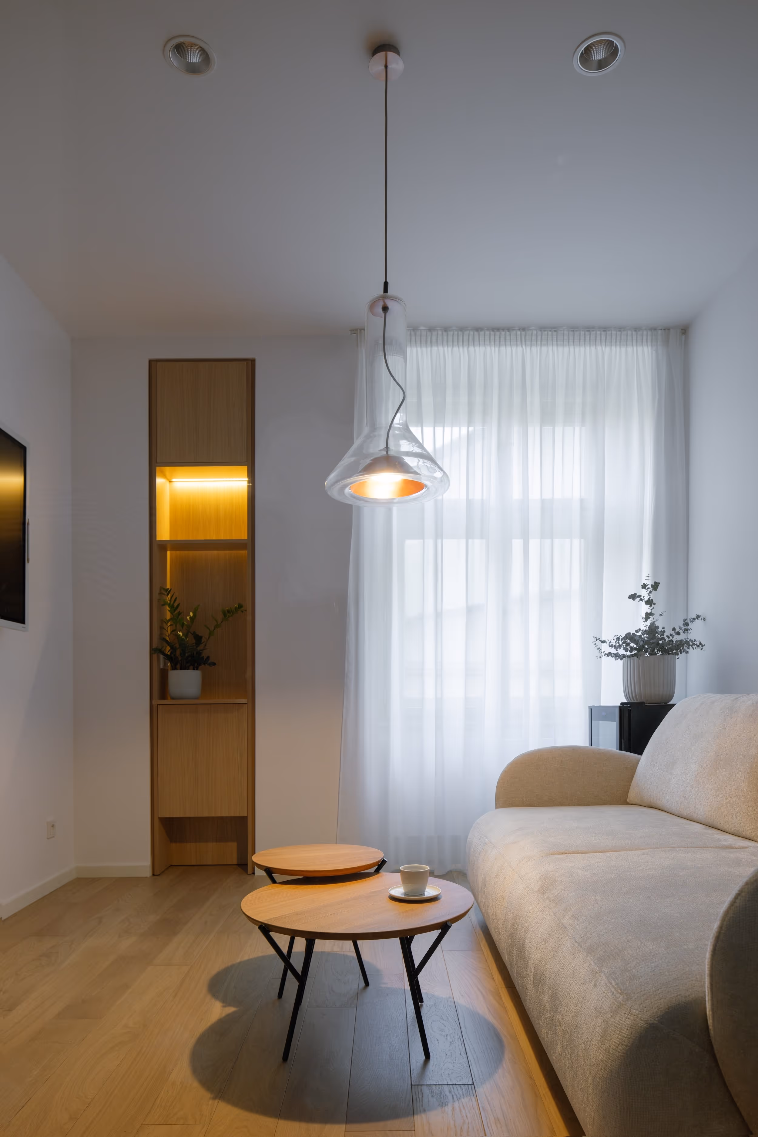 Moderní obývací pokoj s béžovou pohovkou, dvěma dřevěnými konferenčními stolky, visící průhlednou lampou a police s osvětlením a květinou.