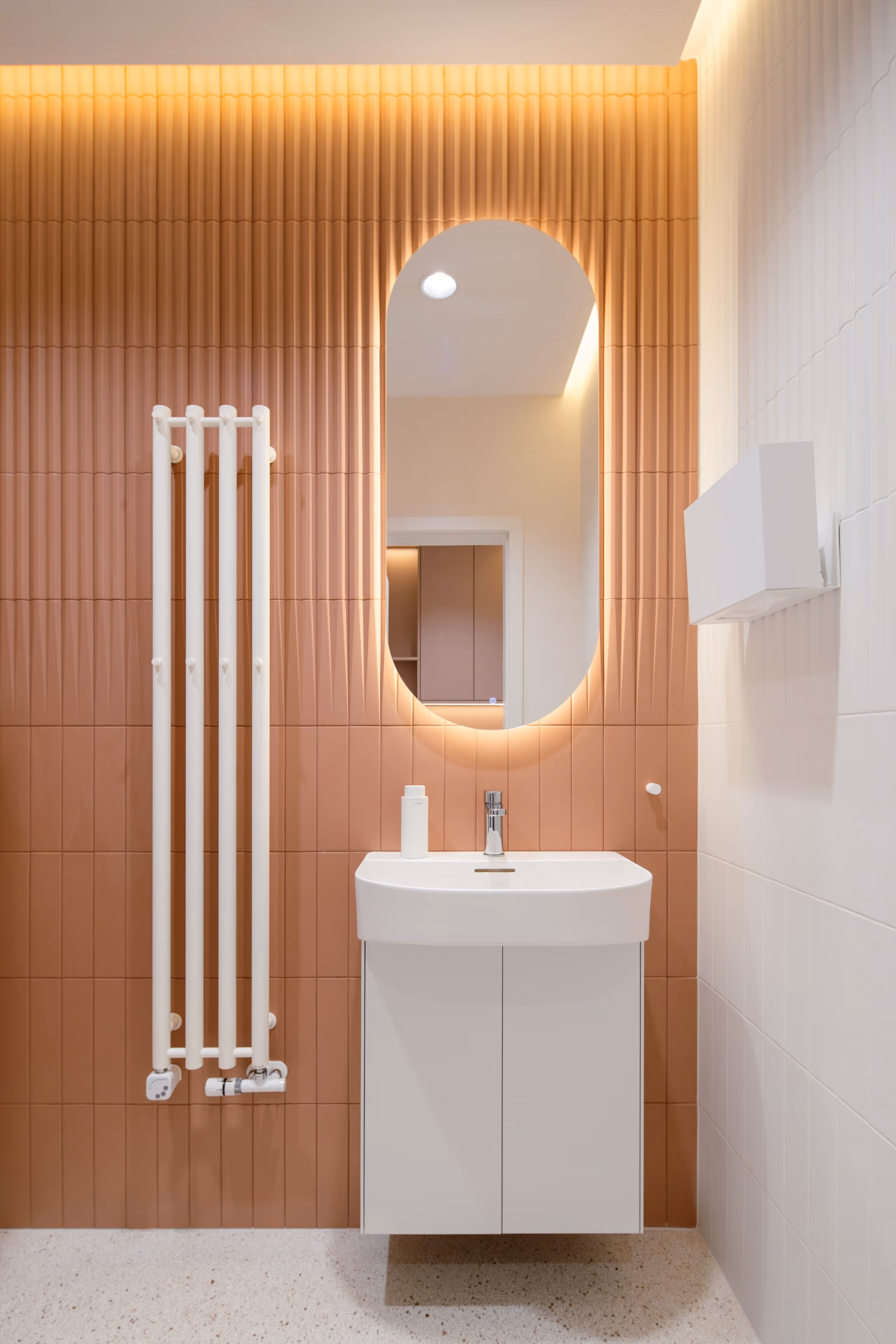 Moderní koupelna s oranžovými obklady, bílým závěsným umyvadlem, zaobleným osvětleným zrcadlem a bílým topením na stěně.