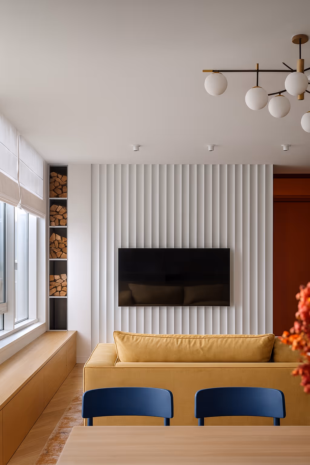 Moderní obývací pokoj s látkovou žlutou pohovkou, závěsnou televizí na bílé stěně s vertikálními lištami a vestavěnými policemi na dřevo vedle okna.