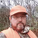Bearded man wearing glasses, an orange cap, and an orange jacket in an outdoor wooded area.