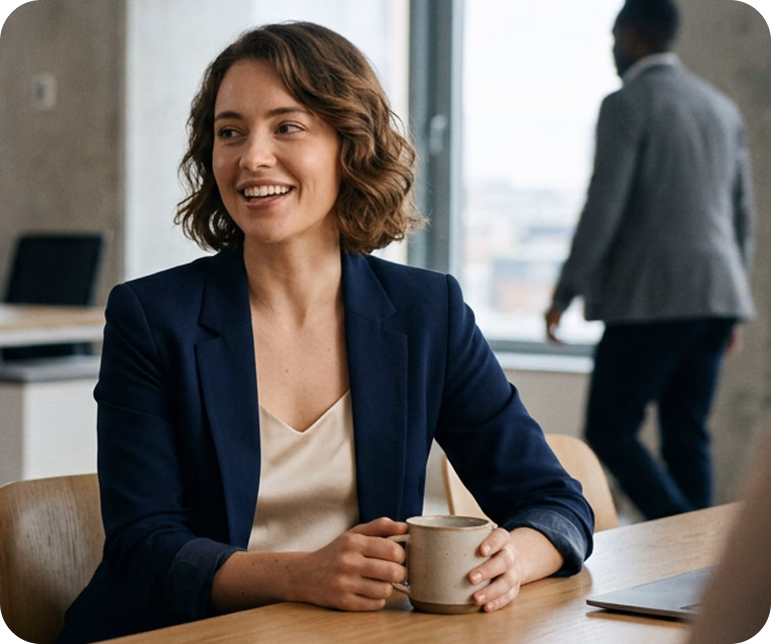 Woman in blazer working on a laptop at office desk with blurred colleagues in background.