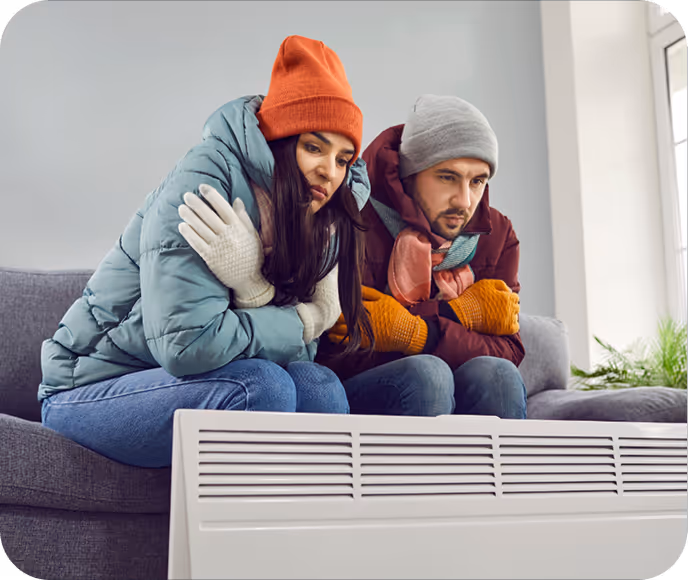 Ein Mann und eine Frau sitzen auf einem Sofa, tragen warme Winterkleidung und wärmen sich vor einem Heizgerät.